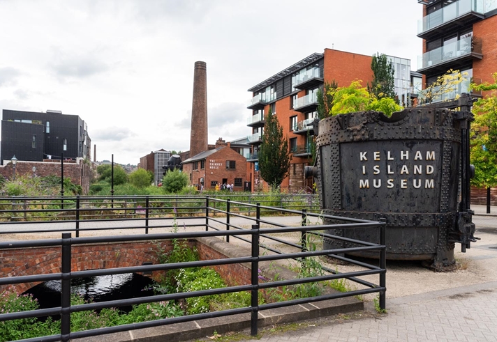Outdoor view of Kelham Island Museum in Sheffield. A large industrial metal container with “Kelham Island Museum” written on it stands in the foreground near railings and a water channel. Surrounding the area are modern apartment buildings, a tall brick chimney, and greenery under a cloudy sky.