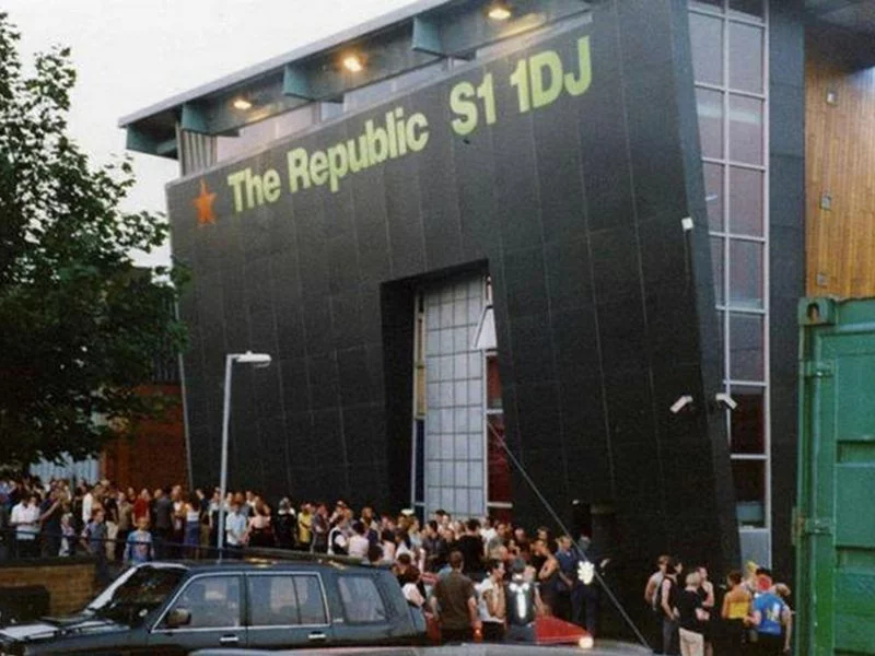 A large modern building with a slanted black facade featuring bold green text that reads “The Republic S1 1DJ” and a red star logo. A crowd of people is gathered outside, forming a long line along the pavement. In the foreground, a dark-colored vehicle is parked near the entrance. The scene appears to be at an event or nightclub, with trees and streetlights visible on the left side.
