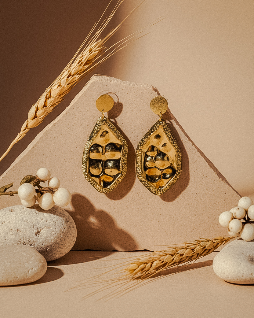 Bold handcrafted earrings with textured gold and black abstract design, displayed against a neutral backdrop with wheat stalks and smooth stones for a natural aesthetic