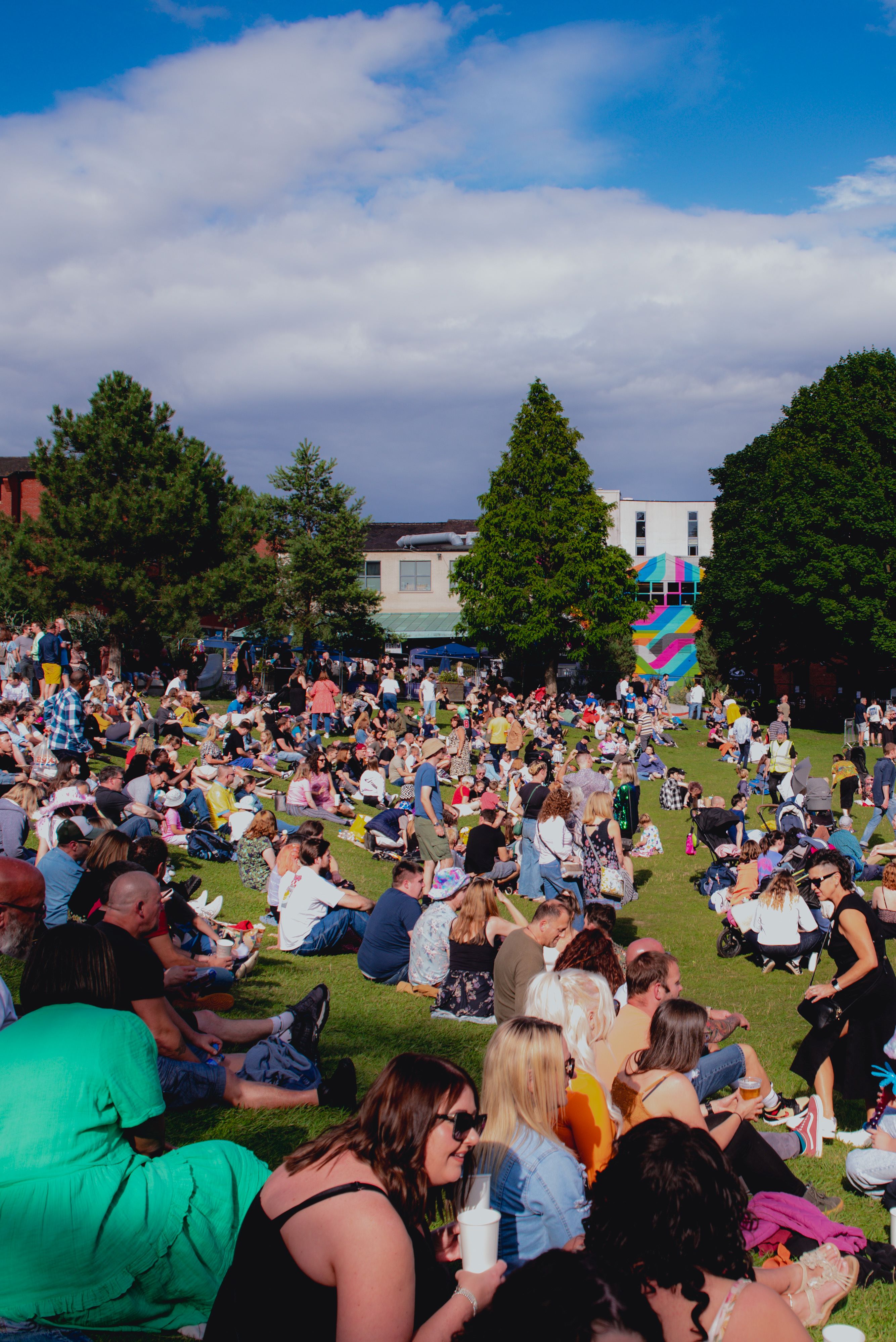 A big crowd of people are sitting on an open are of grass in the sunshine, enjoying an outdoor event in the centre of Sheffield.