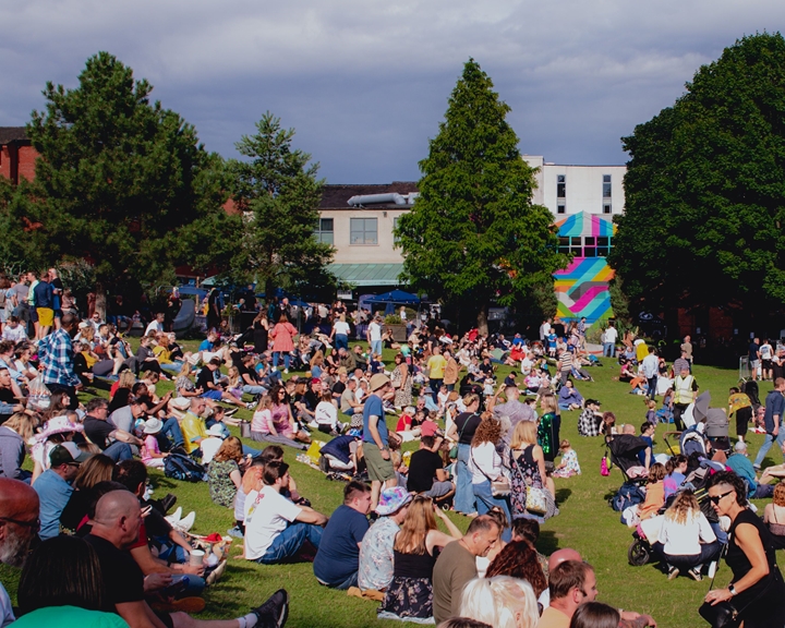 A big crowd of people are sitting on an open are of grass in the sunshine, enjoying an outdoor event in the centre of Sheffield.