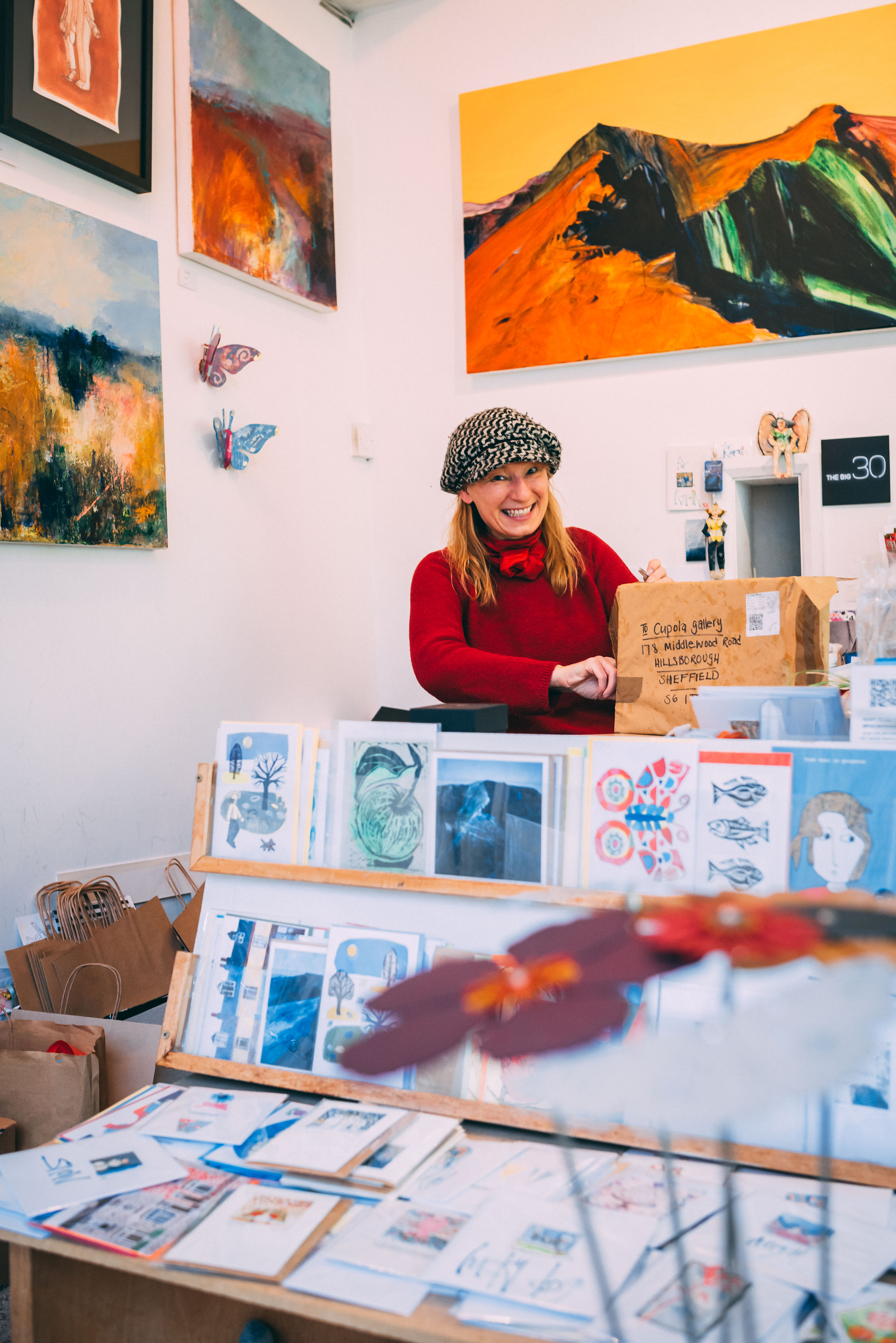 Inside Cupola Gallery at Hillsborough. The walls are covered with paintings by local artists. In the foreground is a display stand filled with prints and greetings cards. A smiling woman is standing in the background opening a large package.