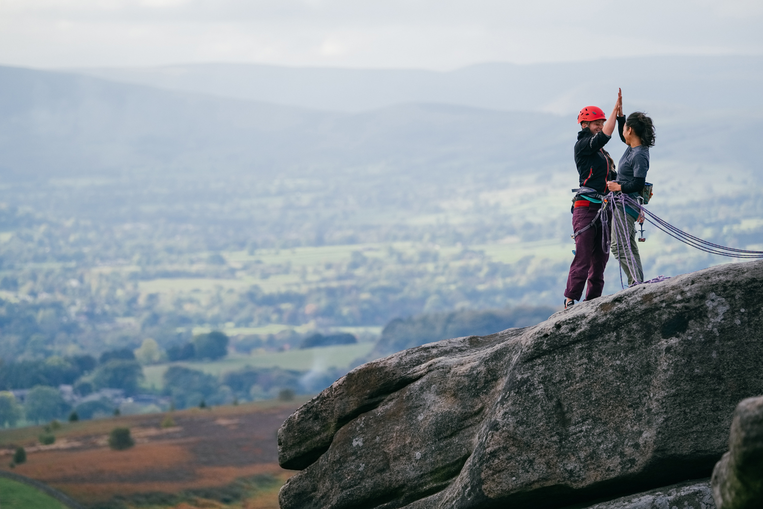 Two people in climbing gear are celebrating on top of a rocky outcrop.