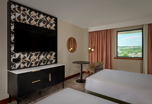 Modern hotel room with two beds, a large flat-screen TV mounted on a patterned panel above a black cabinet, a round mirror on the wall, a small round table with a chair, and a window with pink curtains showing a scenic view outside.