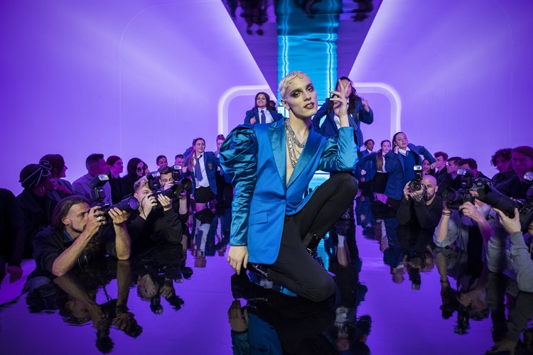 A person in a bold blue outfit with exaggerated shoulders poses confidently on a reflective runway. Purple lighting illuminates the background, while photographers surround the runway, capturing the moment at what appears to be a fashion show.