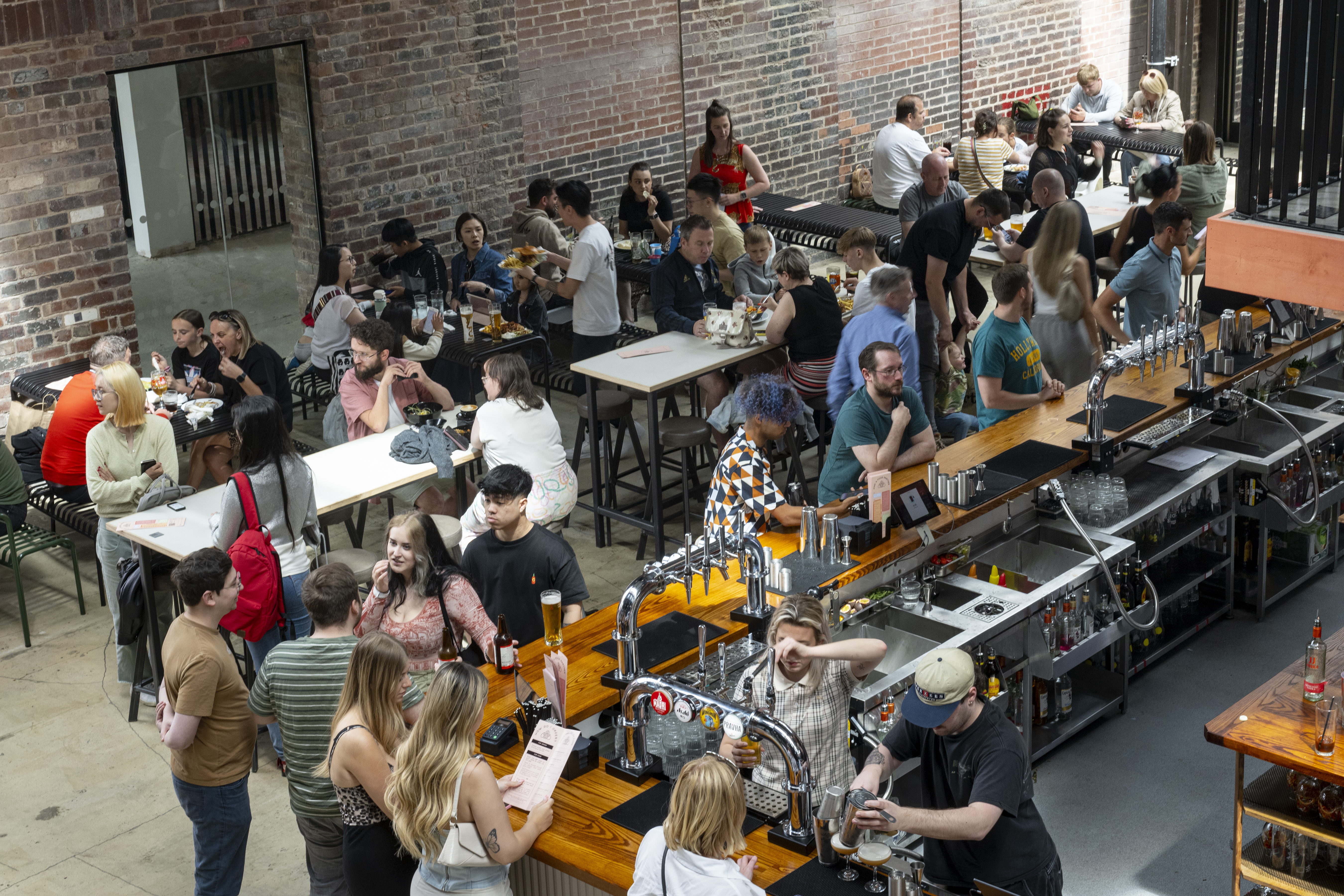 Inside the Cambridge Street Collective food hall there are rows of long tables filled with people eating and chatting. To one side is a bar where people are being served drinks.