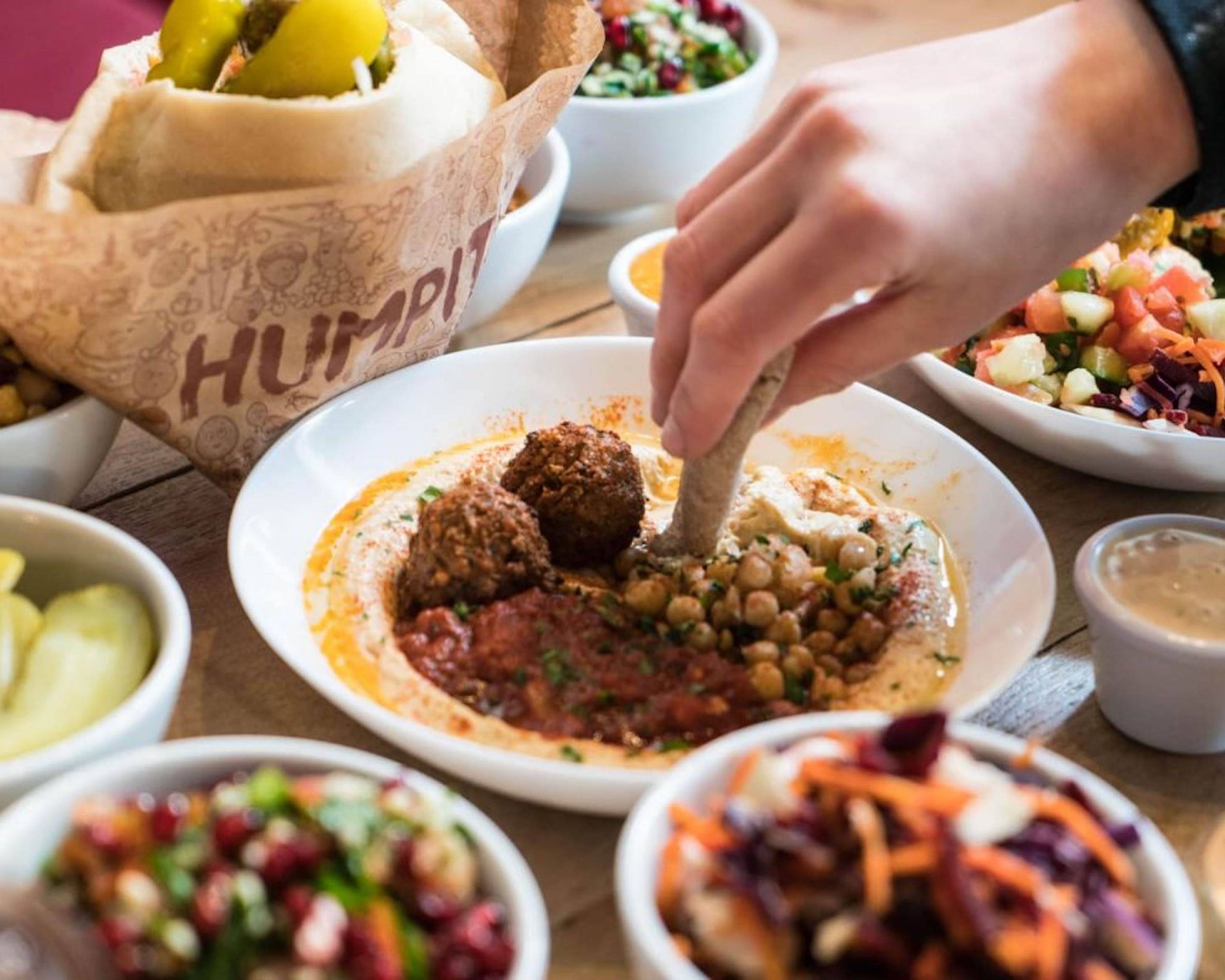 Close-up of a hand dipping pita bread into a plate of hummus topped with chickpeas, falafel, and a red sauce garnish. Surrounding the main dish are several small bowls filled with colorful salads, pickles, and toppings, along with a wrapped pita sandwich in branded paper. The table surface is wooden, and the food presentation emphasizes fresh Mediterranean flavors.