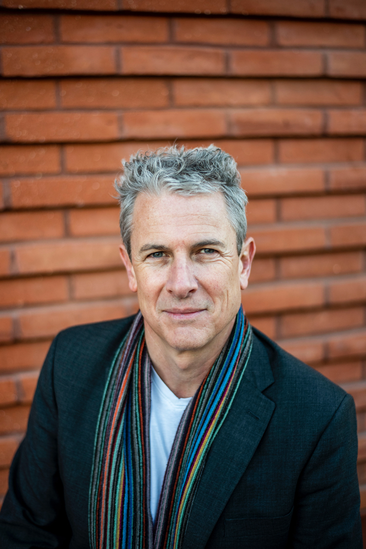 A headshot of Morgan Killick . Person wearing a dark blazer over a white shirt, accessorized with a multicolored striped scarf. The background consists of a wall made of horizontal reddish-brown bricks, creating a textured backdrop. The individual is positioned centrally in the frame, with the upper body visible.