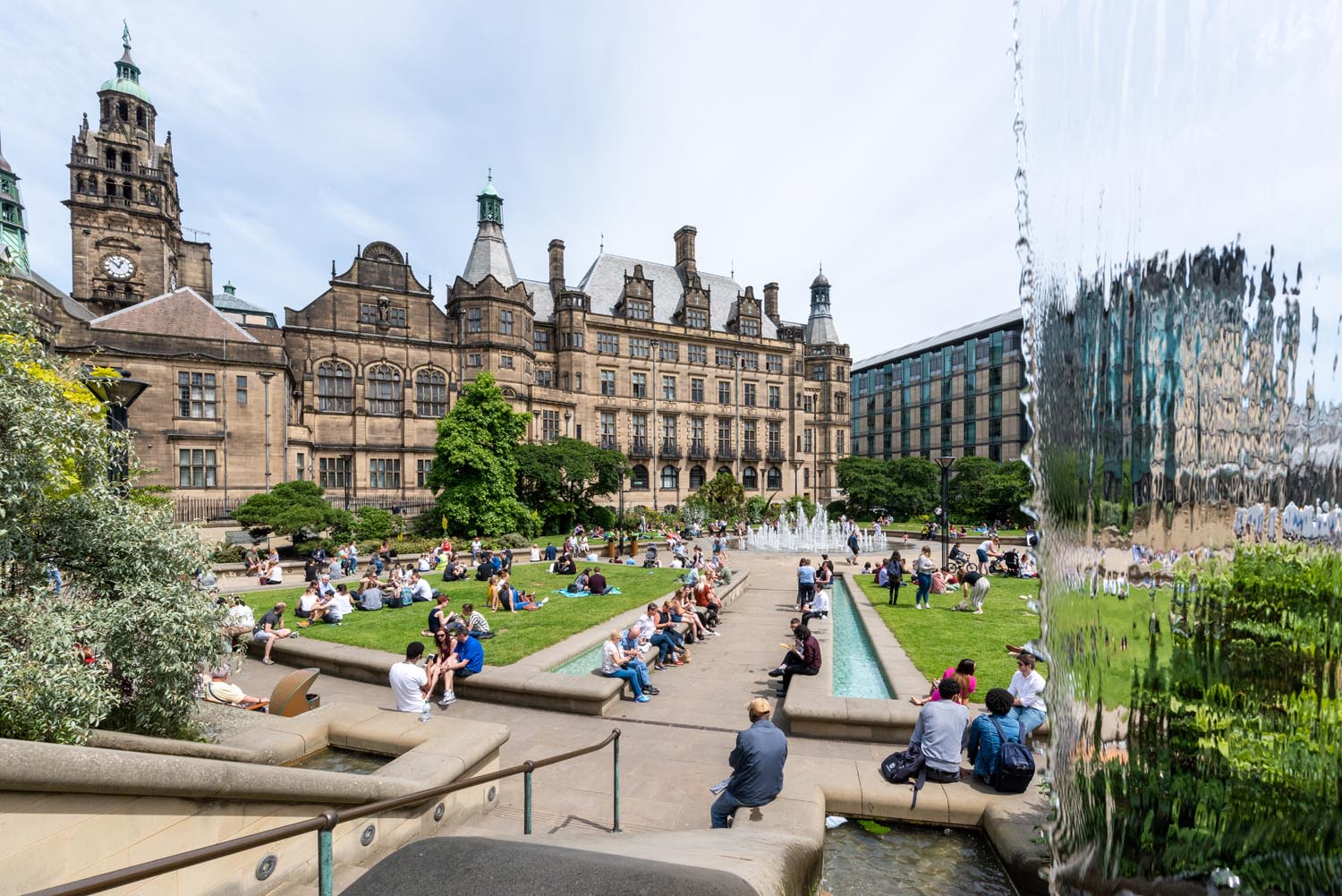 The Peace Gardens, in the centre of Sheffield, on a sunny day. There are lots of people sat on the grass and on benches enjoying the sunshine. In the background is Sheffield Town Hall.