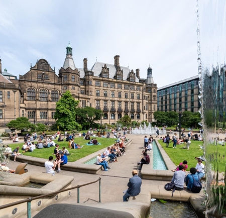 The Peace Gardens, in the centre of Sheffield, on a sunny day. There are lots of people sat on the grass and on benches enjoying the sunshine. In the background is Sheffield Town Hall.
