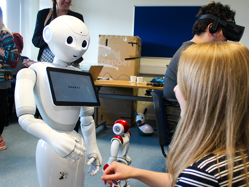 A woman interacting with a humanoid robot which has a touch screen on its chest. There are three other people in the room, one of whom is wearing a virtual reality headset.