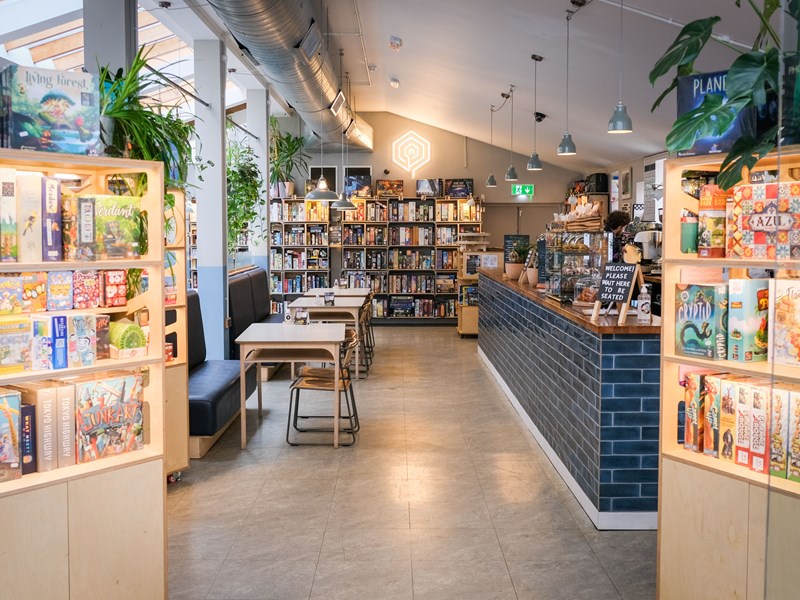 Inside The Treehouse Cafe. You can see tables and chairs, the counter and three shelving units full of board games.