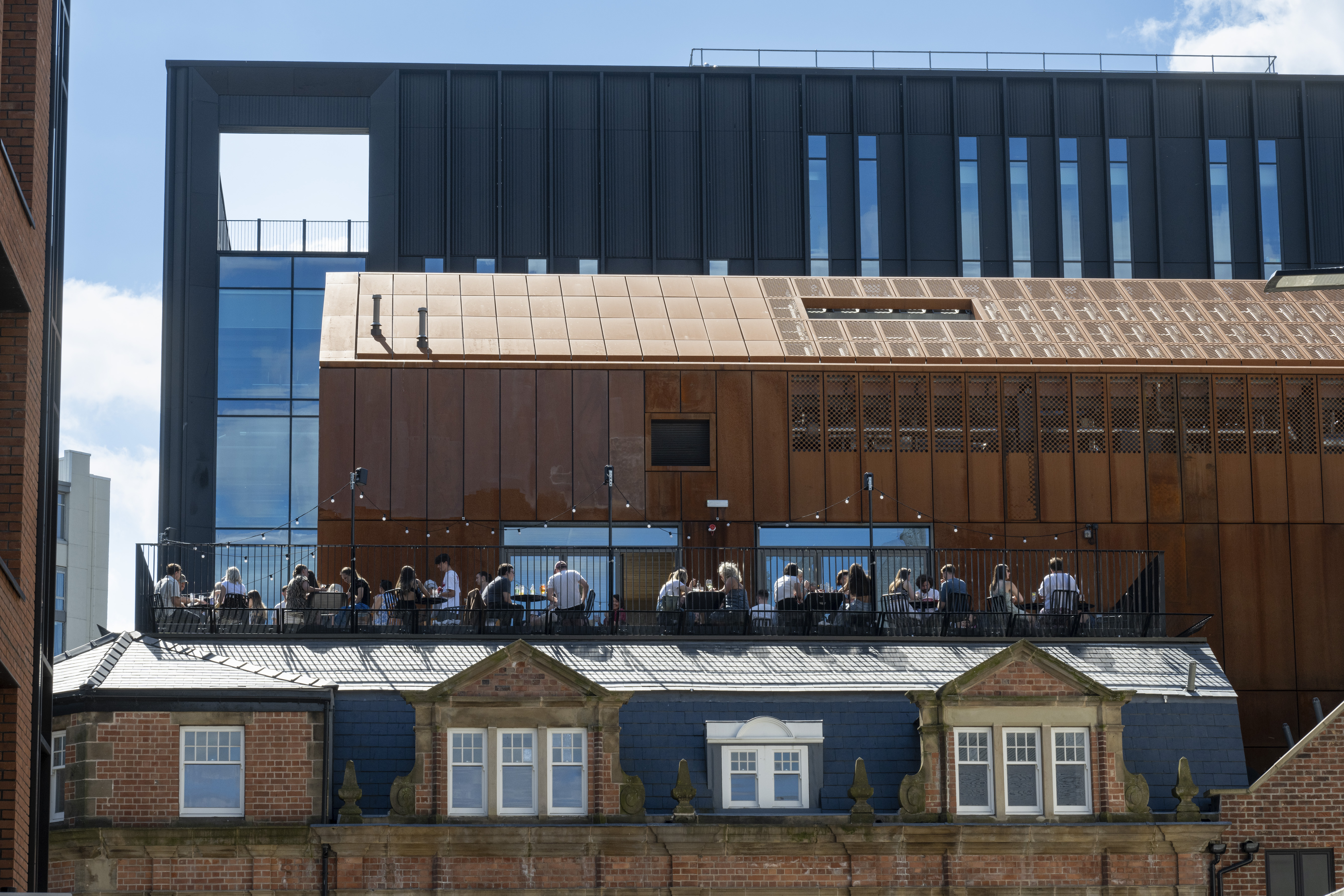 People seated at an outdoor rooftop terrace enjoying food and drinks. The terrace is part of a modern building with a copper-toned facade and large glass windows, set against a backdrop of contemporary architecture featuring black panels and blue-tinted glass. In the foreground, an older brick building with decorative stonework and dormer windows contrasts with the modern structures above.