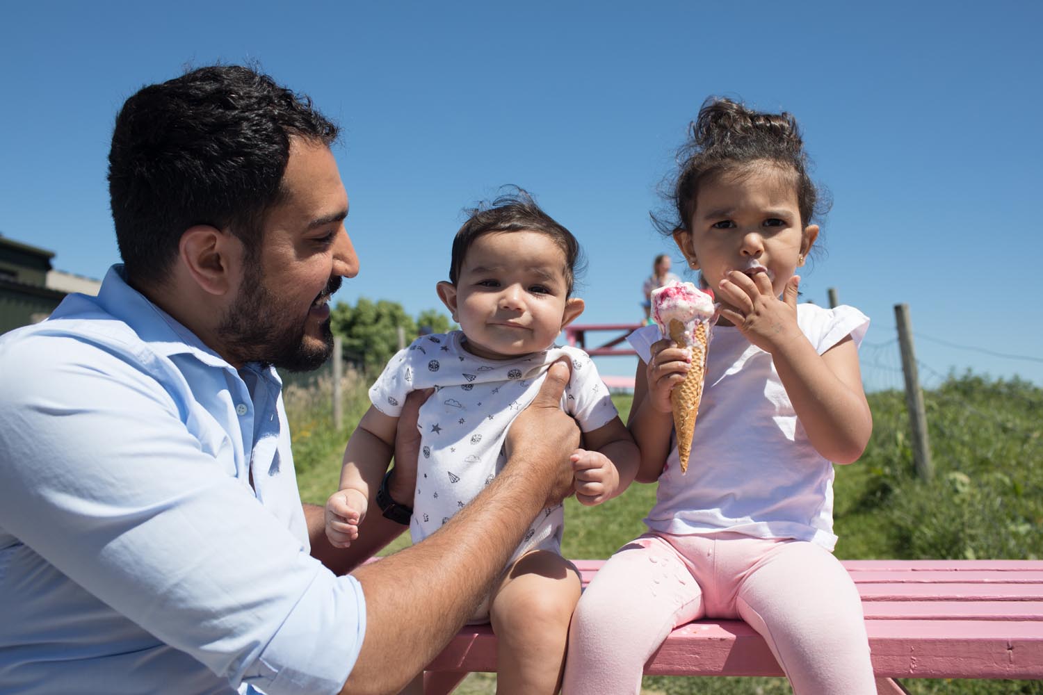 Two small children are sat, with their dad, outside on a bench. The children are eating ice-cream.