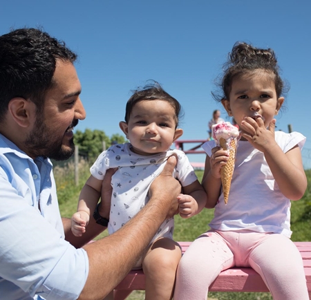 Two small children are sat, with their dad, outside on a bench. The children are eating ice-cream.