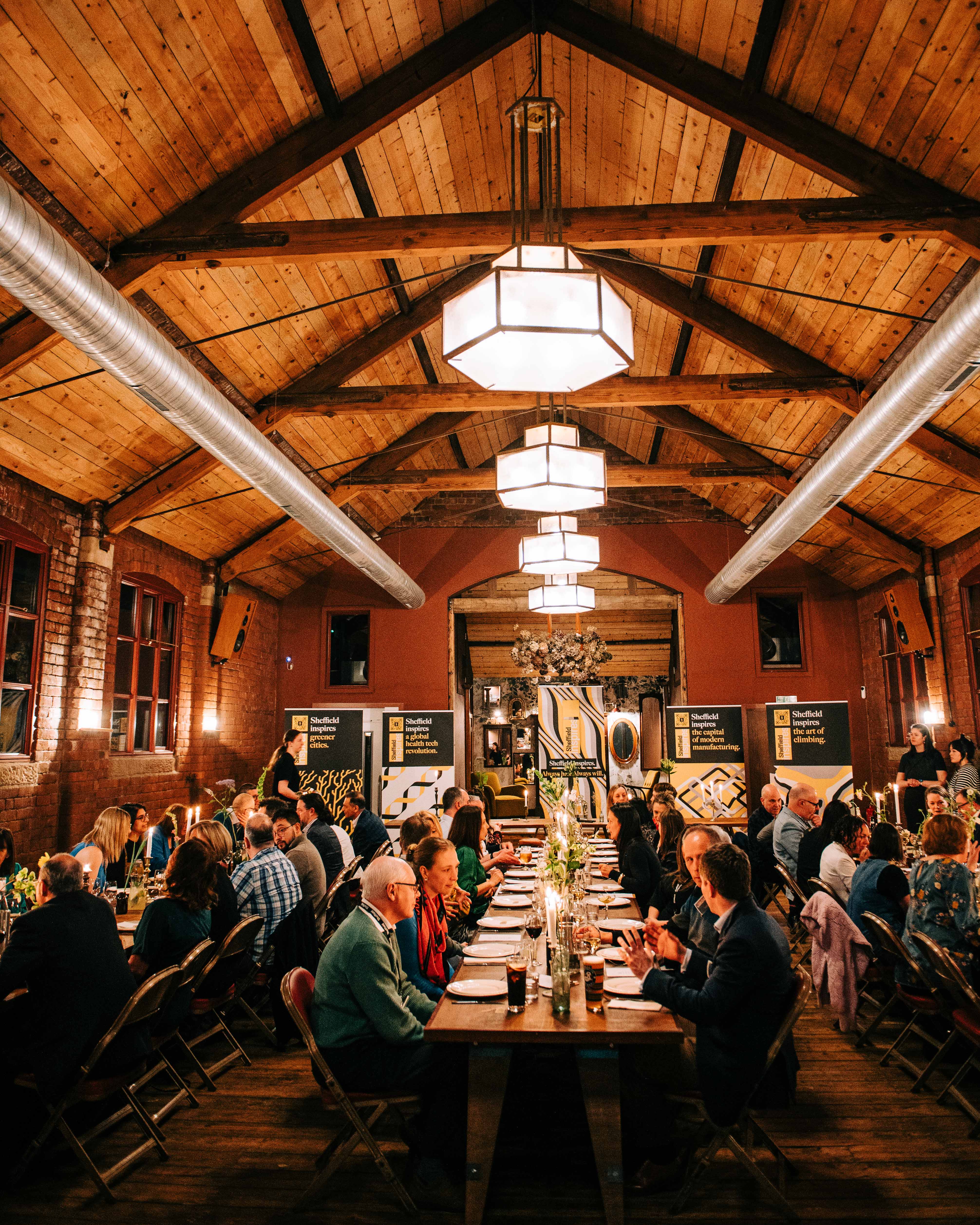A long wooden dining table set for a formal event inside a rustic hall with exposed brick walls and a high wooden-beam ceiling. The room is warmly lit by large geometric pendant lights and decorated with flowers and candles along the table. People are seated on both sides, engaged in conversation. In the background, banners and artwork are displayed near the far wall, adding to the elegant atmosphere.