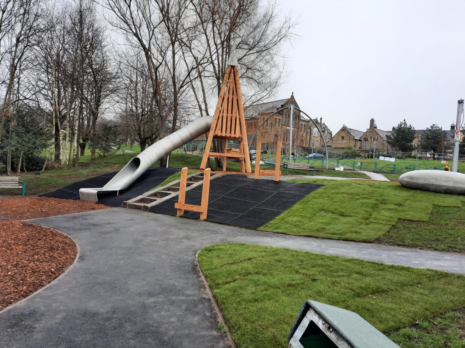 Playground with a large wooden-framed structure and a long, curved metal slide. The area is surrounded by grass, pathways, and leafless trees. Stone buildings and fencing are visible in the background under an overcast sky.