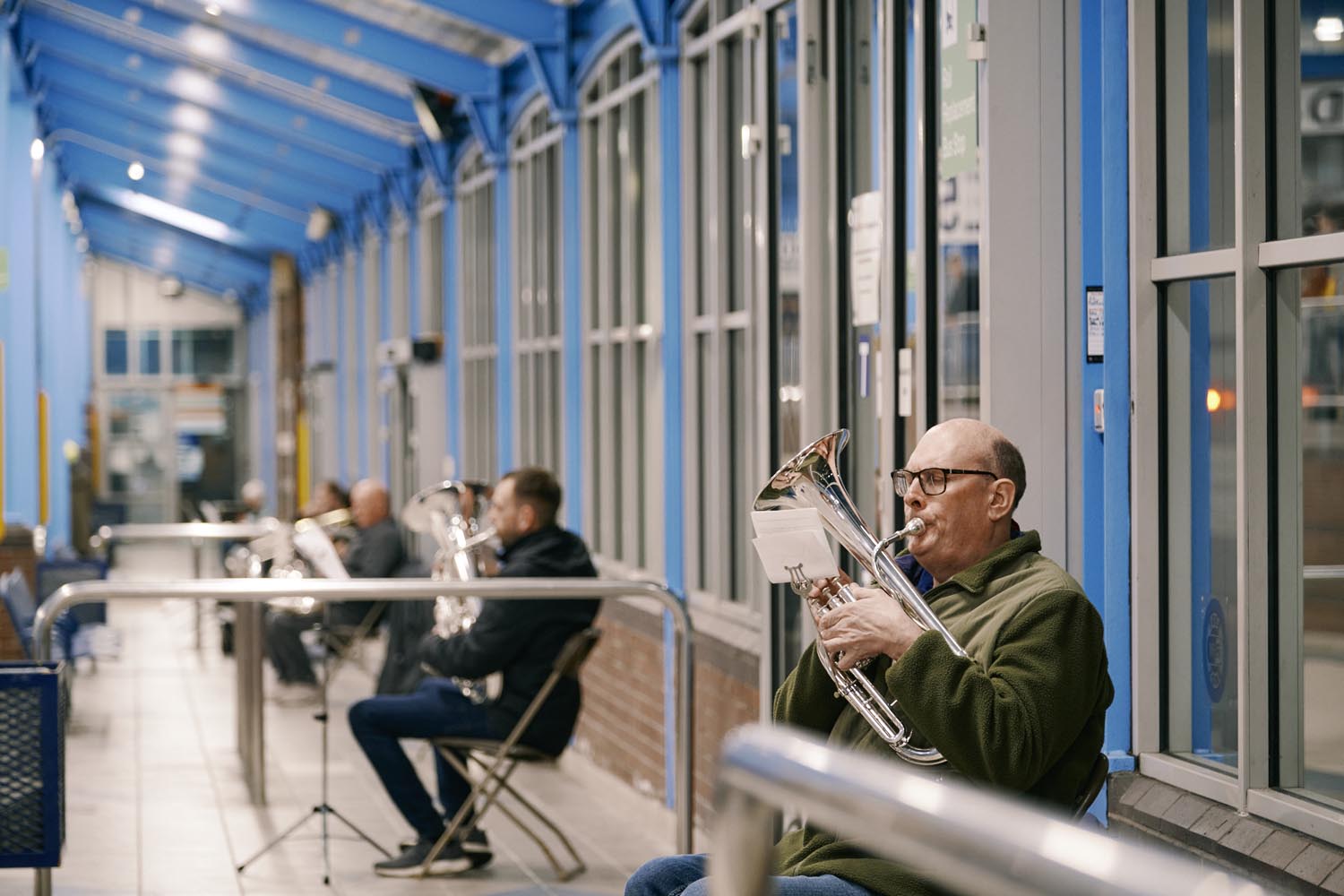 Several musicians seated in a row inside a covered walkway with bright blue metal framework and large windows, playing shiny brass instruments. The setting appears to be an indoor or semi-outdoor public space, with chairs arranged along the wall and instrument stands nearby. The scene captures a casual rehearsal or performance in a modern, well-lit environment.