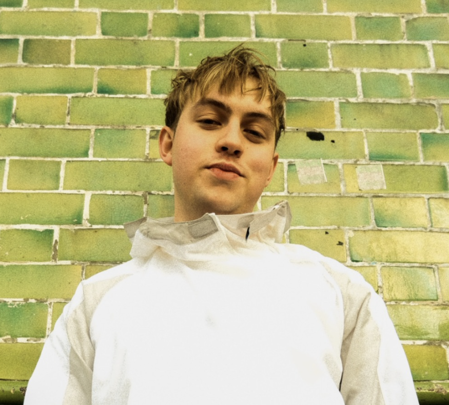 Person wearing a light-coloured jacket standing against a wall made of green bricks with varying shades and weathered texture.