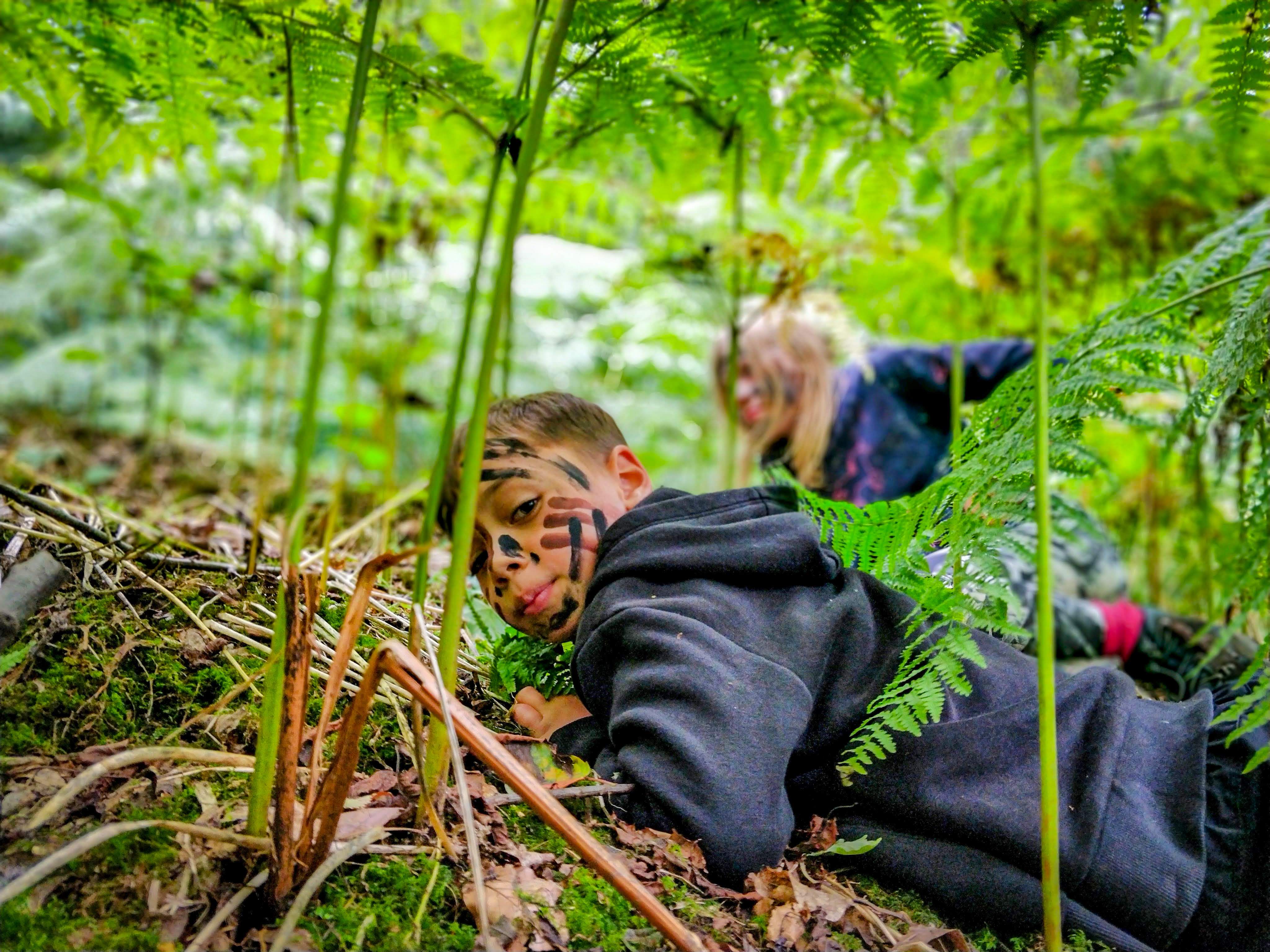 Two children, with face-paint on, are creeping through the undergrowth at TRIBE Bushcraft Centre