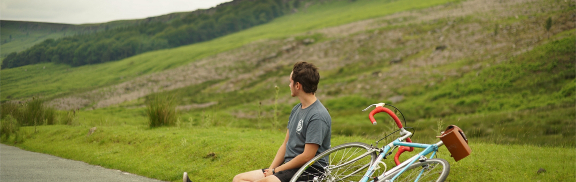 A cyclist sat on the edge of the road having a rest, out in the countryside, with his bike lying on the ground.