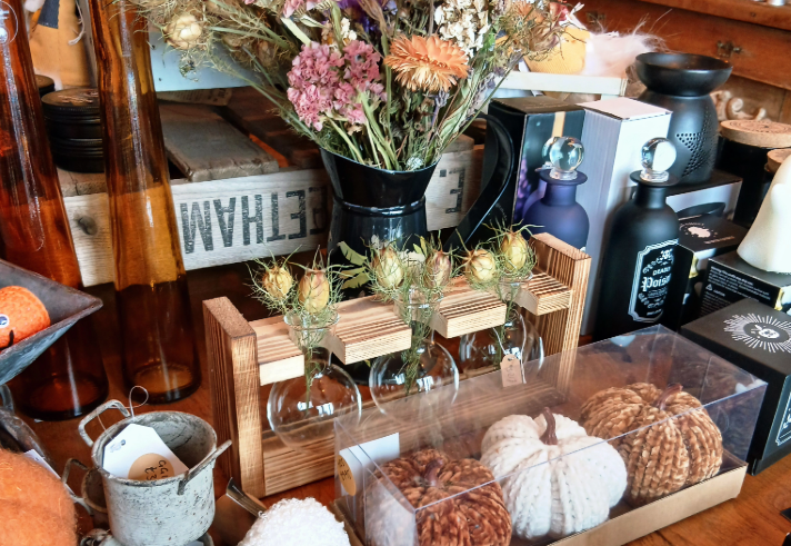 A tabletop display of rustic home décor items, including a black vase filled with dried flowers in shades of pink, purple, and beige, a wooden holder with three small glass vases containing dried thistles, and a clear box with textured decorative pumpkins in white, brown, and tan. Surrounding items include small metal cups, fluffy pom-poms, amber glass bottles, and boxed candles, all arranged in a cozy, autumn-themed setting.