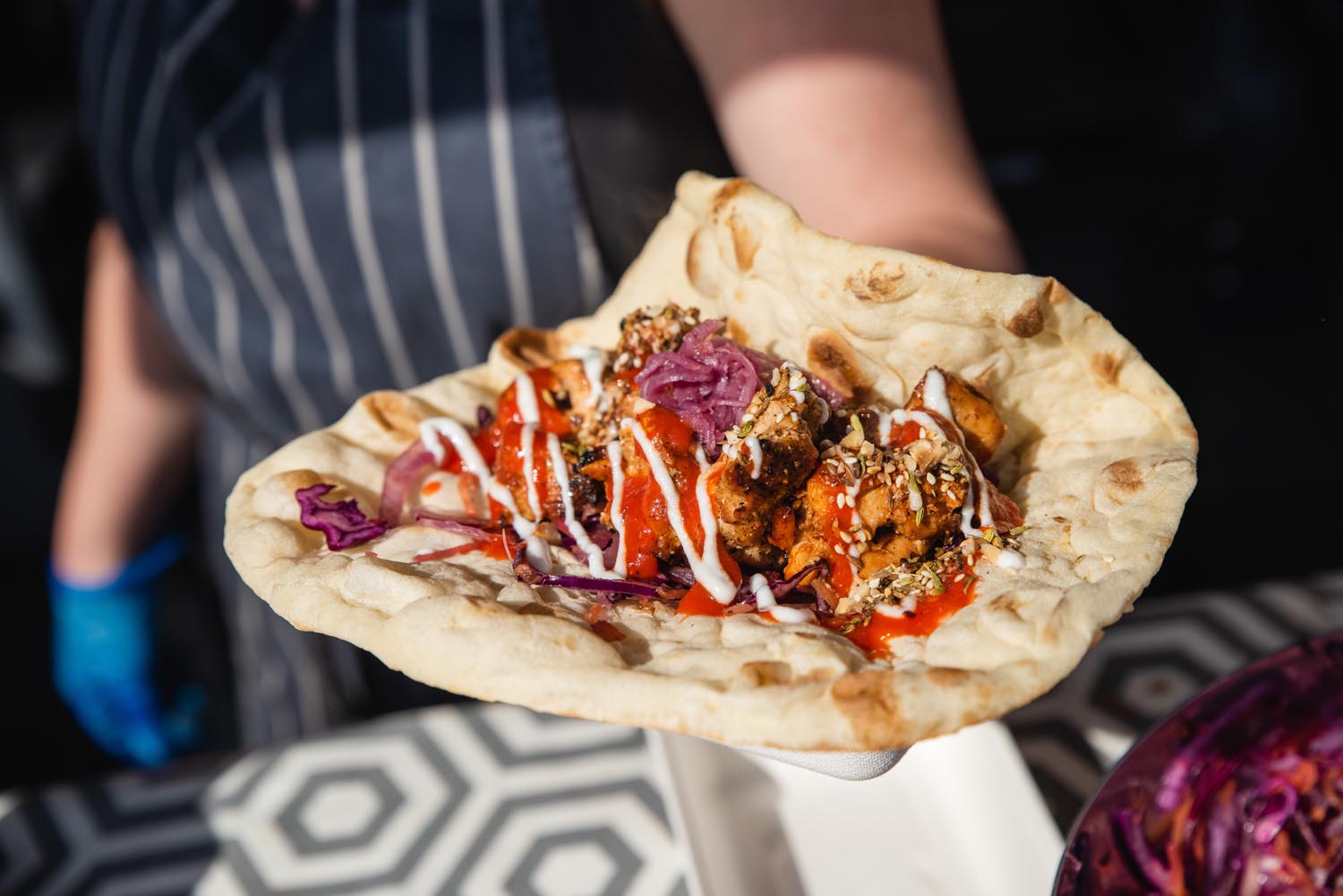 A man in a striped apron holds out a flat bread filled with char-grilled meat, vegetables and sauces.