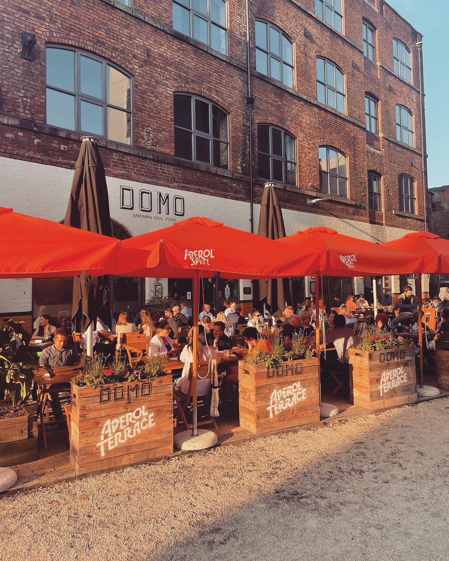An outdoor dining area in front of a brick building with large windows and a sign reading “DOMO.” The seating area is covered by bright red umbrellas labeled “Aperol Spritz” and surrounded by wooden planters filled with greenery. Several people are seated at wooden tables enjoying food and drinks. The scene is lit by warm sunlight, creating a lively and inviting atmosphere.