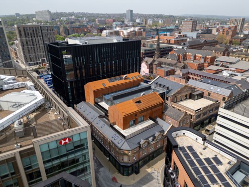 Aerial view of an urban area featuring modern and historic architecture. Prominent buildings include a black glass high-rise and orange-clad structures surrounded by older brick buildings. The scene shows a mix of commercial and residential properties, with solar panels visible on one rooftop and an HSBC building in the foreground. The background includes a sprawling cityscape with trees and hills under a clear sky.