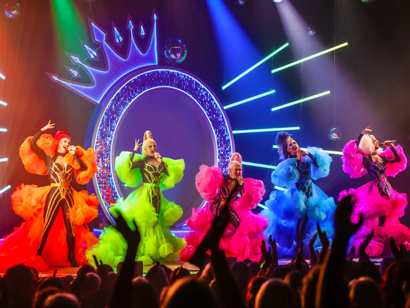 Queenz performing on stage in amazing colourful frocks against a backdrop of a huge neon letter Q with a crown perched on top.