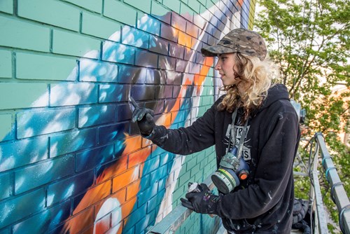 Artist wearing a cap and dark hoodie working on a colorful mural of a bird on a brick wall, using spray paint while standing on a metal platform.