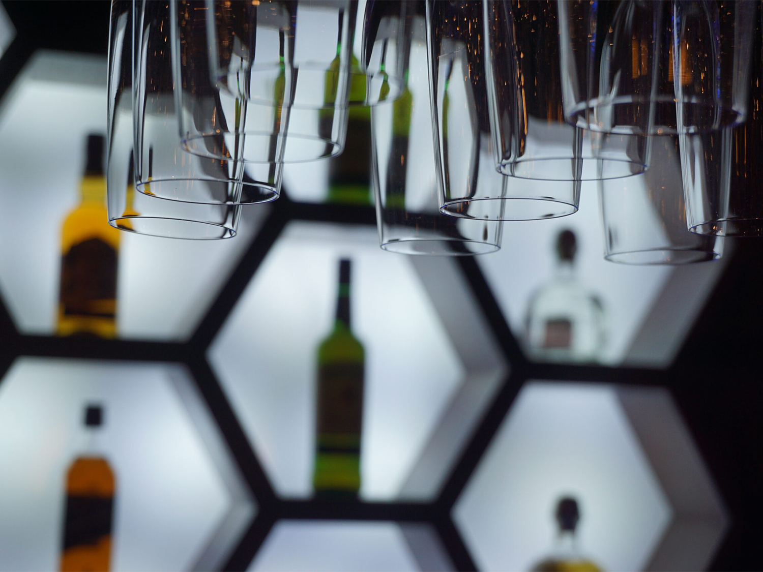Close-up of an illuminated bar display featuring hexagonal shelves with bottles of liquor arranged inside. Several upside-down hanging glasses are in the foreground, creating a layered effect. The shelves have a bright white backlight, contrasting with the dark surroundings and highlighting the shapes of the bottles and glassware.