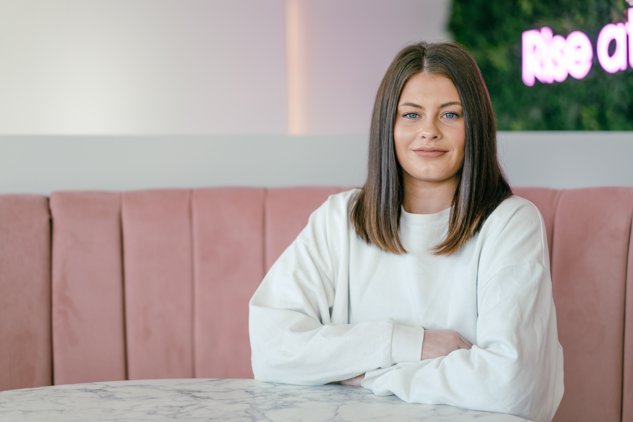 A person wearing a plain white long-sleeve top is seated at a round marble table with arms resting on the surface. The background features a pink cushioned booth and a wall with greenery and partially visible neon text. The setting appears modern and stylish, likely in a café or casual dining space.