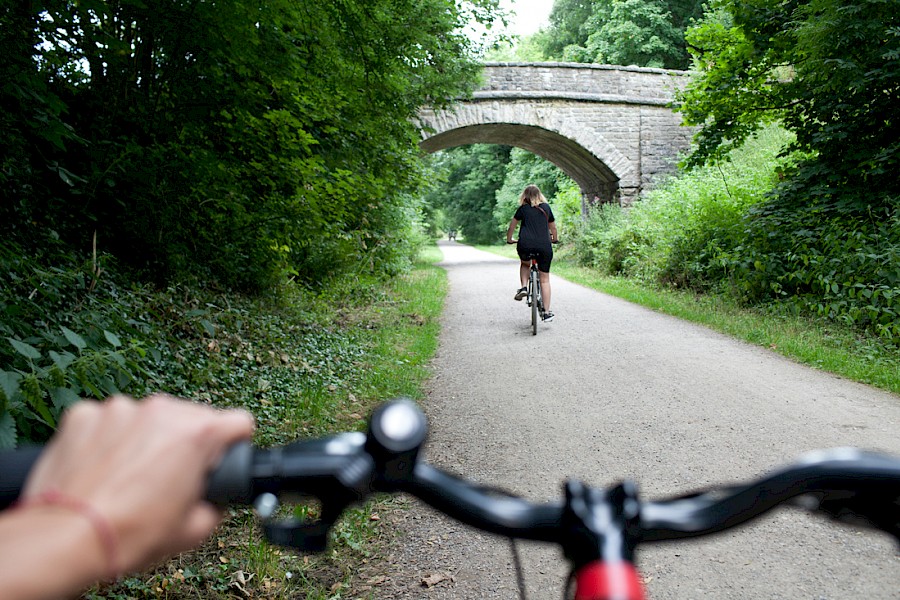 A hand holding the handlebars on their bike, with a cyclist riding along the Monsal Trail in front.