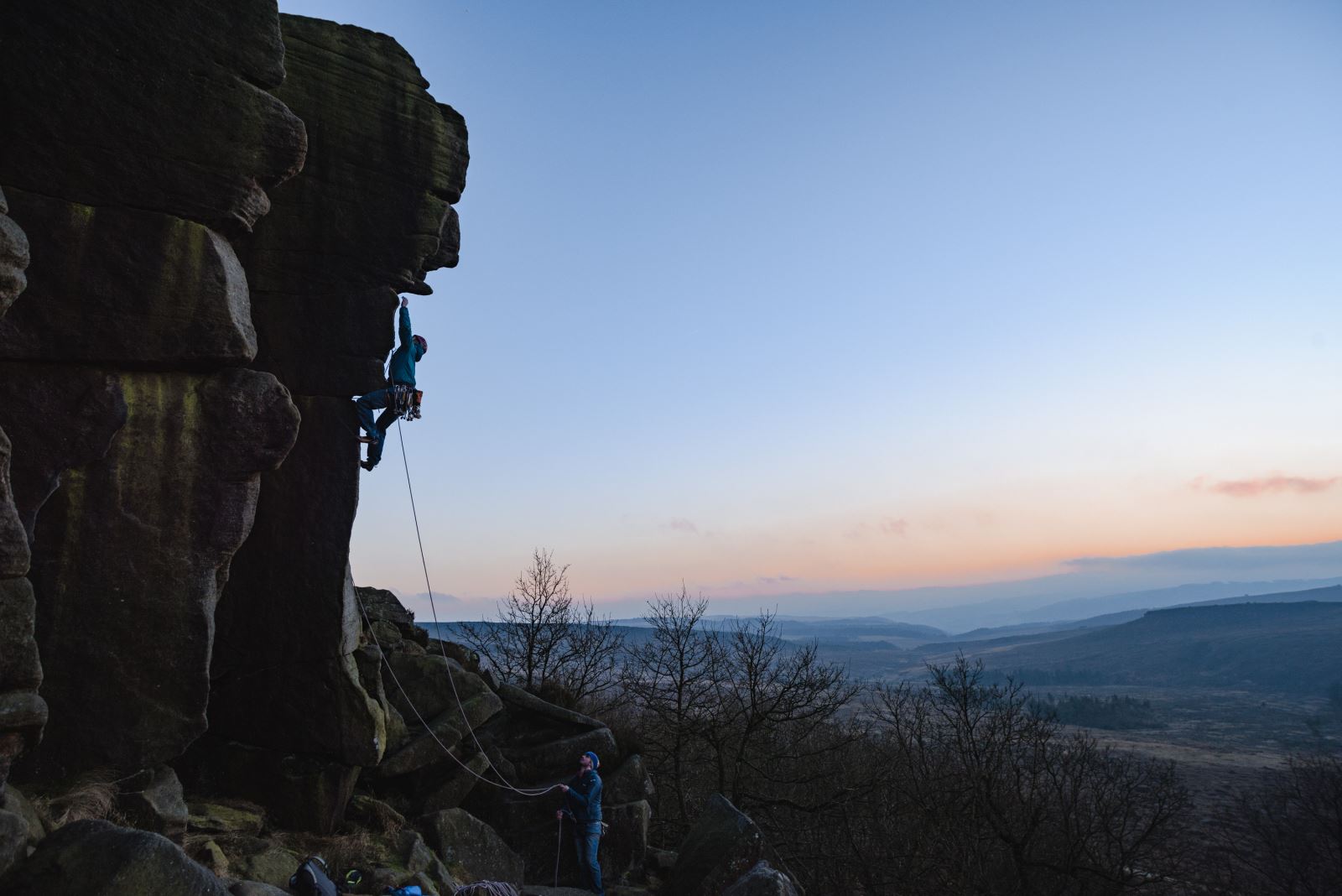 A person is rock climbing on a steep cliff face at dusk, secured by a rope held by another person standing below. The sky is clear with a gradient from blue to orange near the horizon, and the landscape in the background features rolling hills and sparse trees.