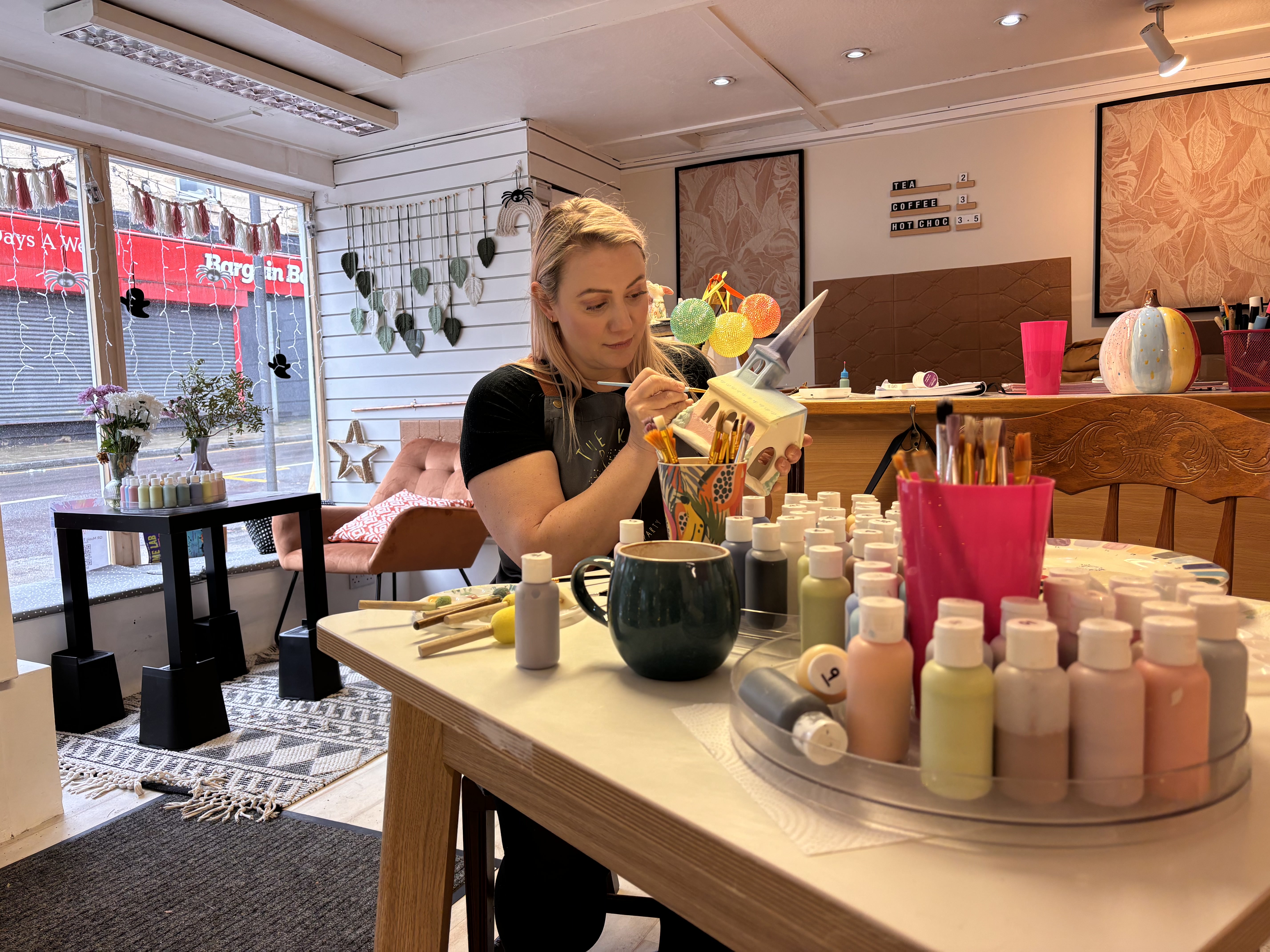 A cozy art studio with a person seated at a table painting a ceramic item. The table is covered with paint bottles, brushes, and a mug. In the background, there are shelves with decorative items, framed artwork on the wall, and a window displaying a street view with a red shop sign. The space is decorated with plants and has warm lighting.