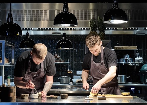 Two chefs wearing dark aprons work at a stainless steel counter in a modern kitchen. One chef is mixing ingredients in a small bowl, while the other arranges food on a wooden board. The background features black tiled walls, hanging black pendant lights, and various kitchen equipment including pots and utensils.
