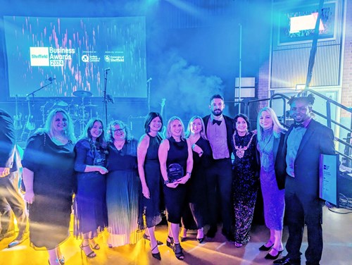 A group of people dressed in formal attire posing together at an indoor event with blue and purple stage lighting. Behind them is a stage with musical instruments and a large screen displaying “Sheffield Business Awards 2023” along with sponsor logos. One person in the center is holding an award, and another on the right is holding a framed certificate. The setting suggests a celebratory awards ceremony.
