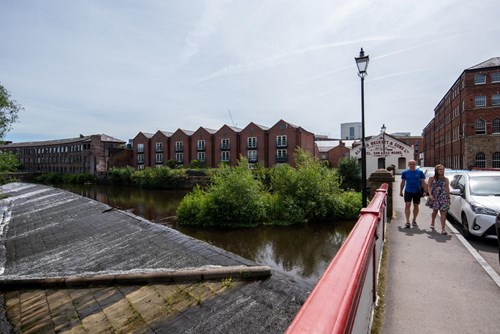 View from a red-railed bridge over a calm river with a small weir on the left. Two people walk along the bridge near parked cars. Across the river are historic red-brick mill buildings with pitched roofs and large windows, surrounded by greenery. A sign on one building reads ‘Kelham Island Museum.’ The sky is clear with light clouds.