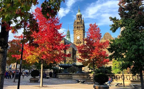 City square with vibrant autumn trees in shades of red and orange, framing a historic building with a tall clock tower in the background. The scene includes ornate fountains, stone balustrades, and people walking around under a bright blue sky with scattered clouds.