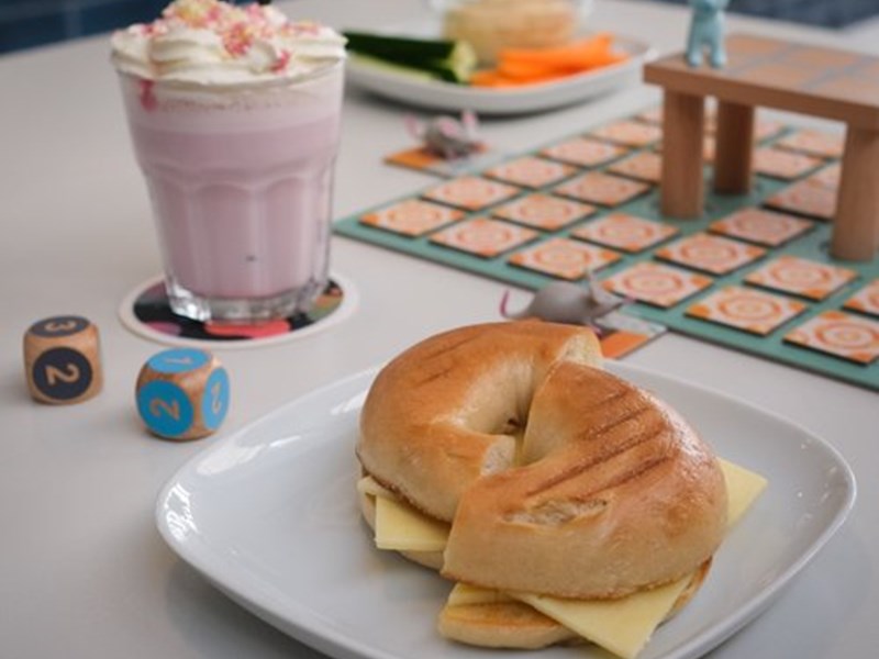 A bagel on a plate and a milkshake, sat on a table that also has a board game set up on it.