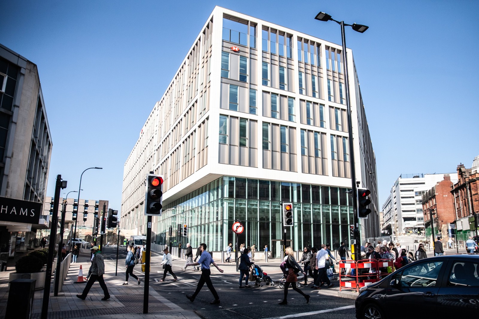 A busy urban street scene with a modern office building featuring large windows and a grid-like facade. Pedestrians cross at a marked crosswalk while cars wait at a traffic light. The clear blue sky and bright sunlight highlight the active city environment.