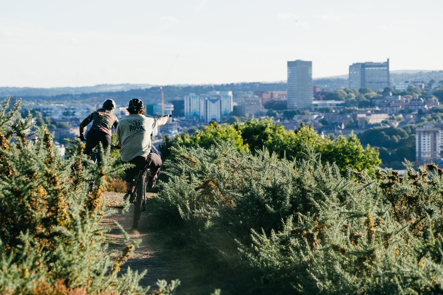 A man and a woman are mountain biking along a dirt track. In the distance you can see Sheffield city centre.