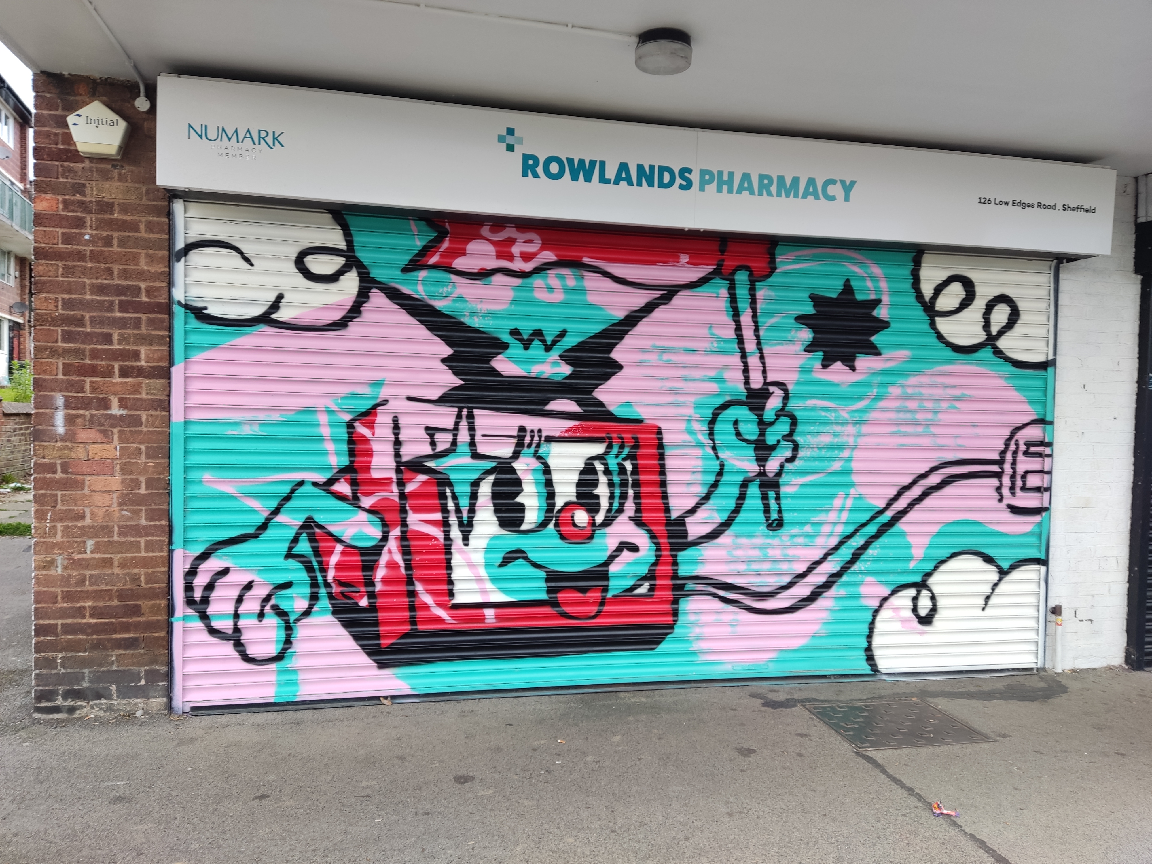 Street art painted on the metal shutter of Rowlands Pharmacy featuring a cartoon-style red and black character with big eyes and a smiling face, holding a wand. The background is turquoise and pink with white clouds and abstract shapes. Above the artwork is a sign reading ‘Rowlands Pharmacy
