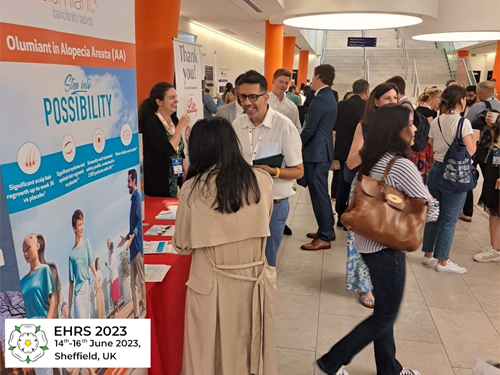 An indoor exhibition area with multiple people interacting at a conference event. A red table in the foreground displays brochures and promotional materials for a medical product related to alopecia areata, with a large banner reading “Olumiant in Alopecia Areata (AA)” and “See the POSSIBILITY.” The background shows other booths, informational posters, and attendees walking or conversing. A sign in the bottom-left corner indicates “EHRS 2023, 14th–16th June 2023, Sheffield, UK.”