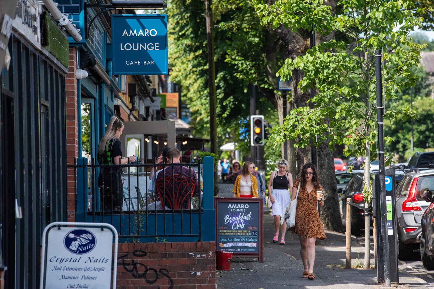 A row of shops and restaurants on a tree lined street. There are people walking up and down the pavement.