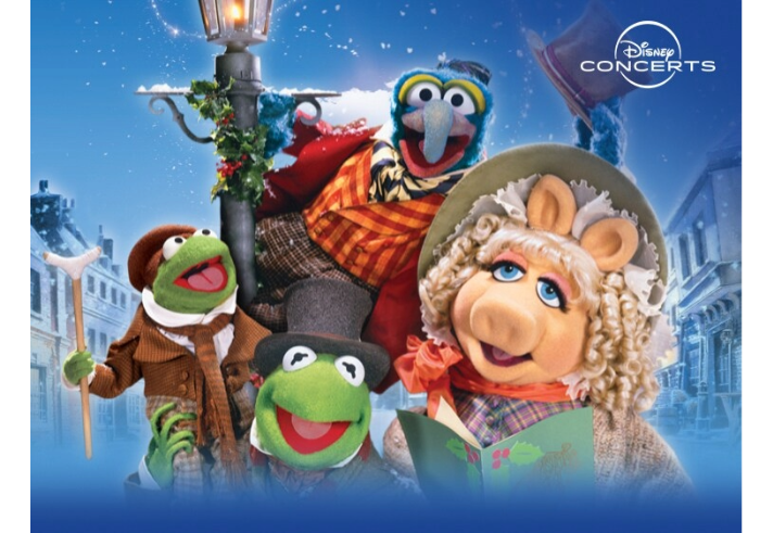 Colorful promotional artwork for Disney Concerts featuring characters from The Muppet Christmas Carol. Kermit the Frog appears twice, dressed in Victorian-style clothing, one holding a walking stick and another wearing a top hat. Gonzo is perched on a lamppost wrapped in festive greenery, wearing a striped scarf and red coat. The background shows a snowy street scene with old-fashioned buildings under a bright blue sky.