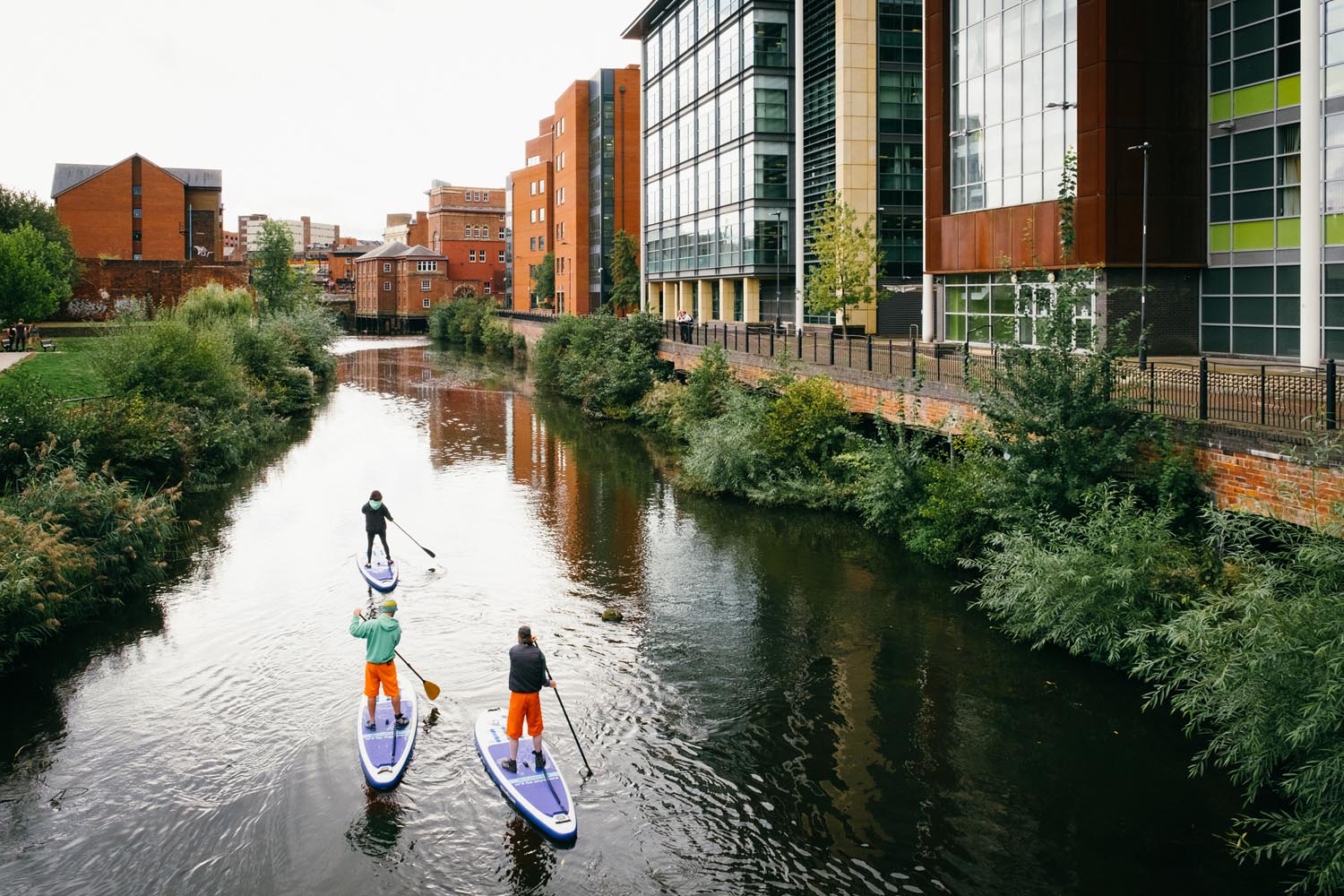 Three people are stand up paddle boarding on a canal that runs through a city centre.