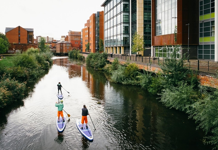 Three people are stand up paddle boarding on a canal that runs through a city centre.
