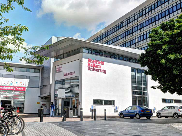 Entrance to Sheffield Hallam University featuring a modern building with large glass windows and white walls. The university's name is displayed in bold red letters. Bicycles are parked on the left, cars on the right, and a tree provides partial shade. People are walking near the entrance under a partly cloudy sky